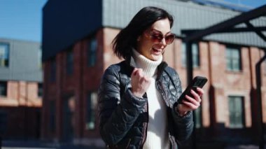 Heyecanlı bir kadın cep telefonunu tutuyor ve yumruğunu sıkarken yüzünü tatmin etmiş bir şekilde duruyor. Şaşırtıcı bayan beklenmedik bir şekilde finansal ikramiye alıyor..