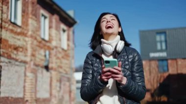 Beyaz kazaklı neşeli kadın dışarıda akıllı telefon ekranına bakarken gülüyor ve kusuyor. Yürümeden önce arkadaşını beklerken kendini komik bir videoyla eğlendiren rahat bir bayan..