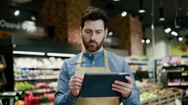 Elinde dijital tablet bulunan önlüklü bir çalışanın portresi ve mağazadaki ürünün durumunu kontrol etmek. Taze meyve ve sebzelerden oluşan sakallı sorumlu adam not alıyor..