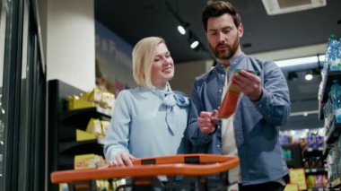 Evli çift, hipermarkette alışveriş arabasının yanında dururken füme balık paketini inceliyor. Bütçeye uyum sağlamak için düşünceli erkek ve kadın ürün fiyatlarını kontrol ediyorlar..