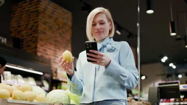 Blondynka Stojąca Wystawie Świeżymi Owocami Trzymająca Ręku Smartfona Cytrynę Uśmiechnięta — Wideo stockowe