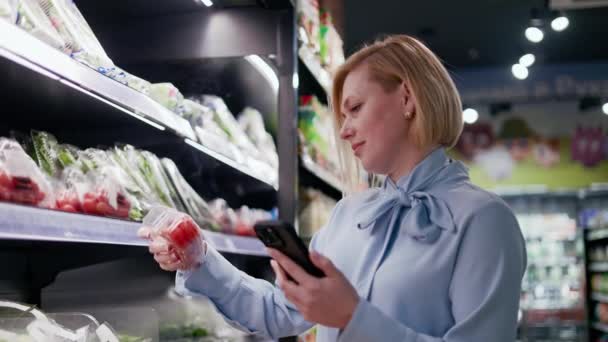 ポータブルスマートフォンを保有しながら 棚からパッケージ製品を選択する若い女性 さまざまな野菜やフルーツを備えた棚付きの明るいブルーシャツを着用している女性 — ストック動画