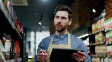 Önlüklü beyaz eşya yöneticisi dijital tablet tutuyor ve satılık malların bulunabilirliğine dikkat çekiyor. Gülümseyen perakende işçisi modern teknolojiyi kullanarak market çeşitliliğini özetliyor..