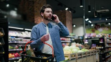 Markette elinde alışveriş sepeti olan yakışıklı beyaz bir erkek modern akıllı telefon konuşmaları yapıyor. Gıda ürünleri alırken internet üzerinden iş sorunları çözen genç sakallı adam..