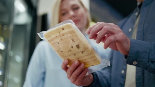 两个人组成的家庭在杂货店里密切注视着切碎的奶酪包装 笑着看着穿着蓝色衬衫的男人 并同意人们对奶制品的看法 — 图库视频影像
