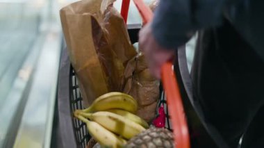 Alışveriş arabasının kapalı mekanda insan eliyle tutulan farklı bakkalların olduğu yakın plan. Kahverengi kağıt torbalar ve taze meyveler. Muz ve ananas da dahil. Market ve süpermarket kavramı.