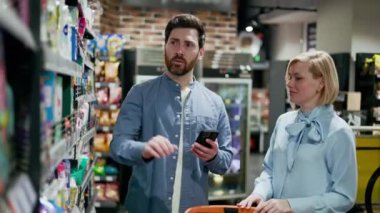 Mağazada iki kişi, çeşitli ürünlerle dolu raflarla koridorda dururken ürünü inceliyor. Yakışıklı bir adam beyaz bir şişe tutuyor ve beyaz bir ortağın yanında inceliyor..