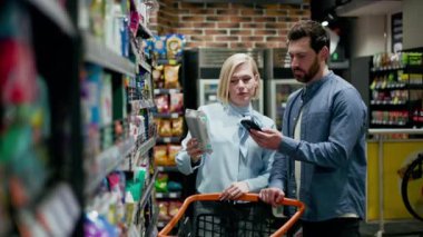 Genç beyaz koca alışveriş arabasının yanında dururken çekici eş marketteki raflardan paket alıyor. Sıradan giyinmiş bir çift. Aperatifler, renkli çantalar ve kutularla dolu koridor..