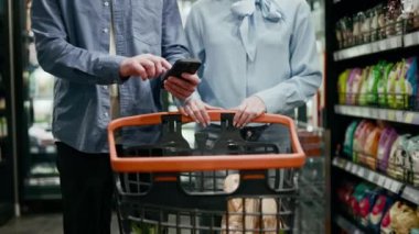 Markette iki kişi. Beyaz bir erkek akıllı telefon tutarken genç bir kadın yanına gidip birkaç eşya ile dolu alışveriş arabalarını itiyor. Satın alma, teknoloji ve alışveriş kavramı.