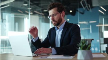 Masada oturan ve modern ofis ortamında taşınabilir laptop üzerinde çalışan hayal kırıklığına uğramış bir erkek. Kapalı alanda baş ağrısı çeken koyu renk takım elbiseli yakışıklı beyaz adam. Acı ve insan kavramı.