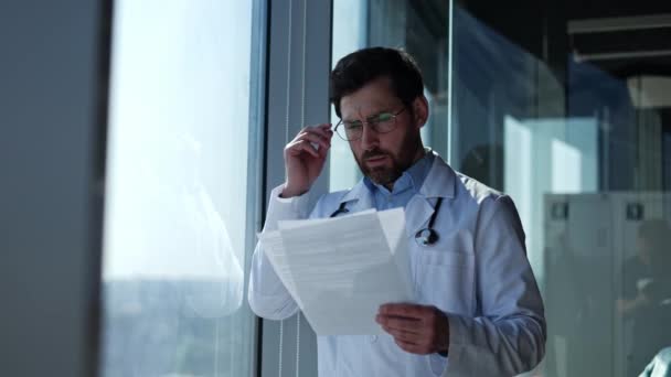忧心忡忡时 担心穿着白衣的男性肿瘤学家站在窗边阅读测试结果 医务工作者摘下眼镜时感到失望 因为他们没有得到任何帮助 — 图库视频影像