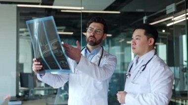 Yetenekli beyaz sakallı erkek röntgenini tutuyor ve parmaklarıyla işaret ediyor. Bu sırada yetenekli Asyalı hastane çalışanı meslektaşlarını dikkatle dinliyor. Sonuç ve karar kavramı.
