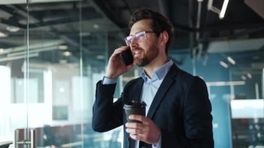 Ofis ortamında kağıt bardak kahveyle akıllı telefon görüşmesi yapan aktif bir şirket çalışanı. Olumlu beyaz adam, kapalı mekanda duygusal uzaktan konuşma için modern aletleri kullanıyor..
