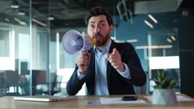 Harika bir iş adamı megafonla bağırıyor ve masanın başında tek başına otururken hareketler yapıyor. İş yerinde kamerayla konuşurken yaratıcı girişimci el sallıyor ve tuhaf bir aksesuar kullanıyor..