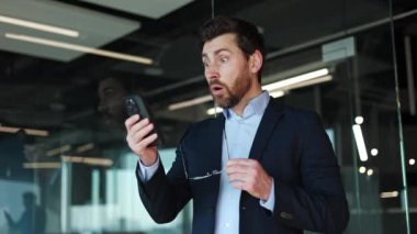 Modern akıllı telefondan haber okurken beyaz bir erkeğin şok geçirmesinin yan görüntüsü. Yakışıklı bir iş adamı, elinde bardaklarla modern ofiste şaşkınlığını dile getiriyor..