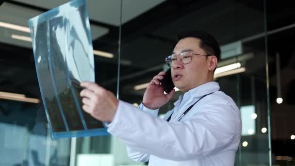 戴眼镜的亚洲外科医生正在检查X光照片 并在手机上交谈 医学专家在研究结果并通过电话咨询同事后得出结论 — 图库视频影像