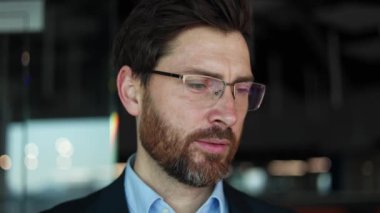 Kapalı alanda kameraya bakarken dişleriyle gülümseyen orta yaşlı erkek portresi. Koyu renk ceketli, mavi yakalı ve arka planda cam gözlüklü yakışıklı bir adam..