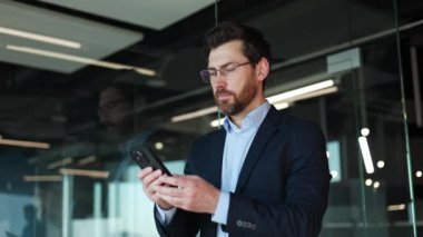 Kafkasyalı bir iş adamı modern aletleri kullanarak geniş camsı ofislerdeki sosyal medyaları kaydırıyor. Ciddi sakallı erkek gözlüğünü çıkarıyor ve kapalı alandaki kişisel akıllı telefon ekranına bakıyor..