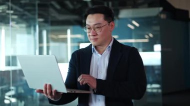Tatmin olmuş Japon erkek dizüstü bilgisayarını elinde tutuyor ve geniş ofis odasında dikilirken etrafı kolaçan ediyor. İş adamı gözlüklü ve takım elbiseli, internetteki işini bitirdikten sonra kameraya bakıp gülümsüyor..