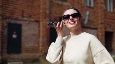 Gayri resmi giyinmiş neşeli bir kadın sokakta yürüyor ve akıllı telefondan sesli mesaj dinliyor. Dışarıdaki arkadaşlarla iletişim kurmak için modern akıllı telefon ve internet bağlantısı kullanan bir kadın..