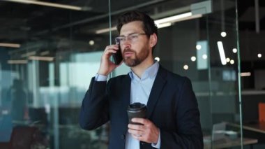Duygusal ofis çalışanı kapalı alanda çalışanlarla konuşmak için kişisel akıllı telefonu kullanıyor. Erkek el kol hareketi yaparken, kağıt bardağı lezzetli bir içecekle tutuyor. Ayrılma ve konuşma kavramı.