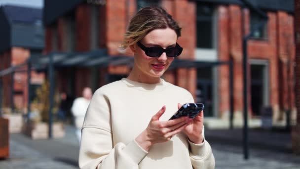 穿着米色运动衫的漂亮女士站在外面看着智能手机屏幕和打字信息 在等公共汽车的时候 用智能手机给女人们戴着凉爽的太阳镜笑 — 图库视频影像