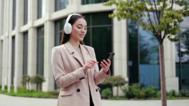 Kulaklıklı beyaz bir kadın modern binanın önünde dururken telefonla şarkı seçiyor. Resmi giyinmiş keyifli kadın halka açık yerlerde müzik zevki için cihazlar kullanıyor..