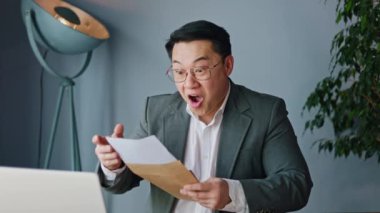 Neşeli Asyalı adam, kişisel ofisteki modern dizüstü bilgisayarla masada otururken hevesle zarfı açıyor ve iyi haberlerle belgeleri topluyor. İçeride neşeli duygular ifade eden keyifli genç adam..