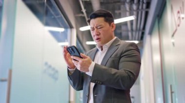 Tasmasını tutan endişeli Japon erkek ofis koridorunda dikilirken kişisel akıllı telefon ekranına bakıyor. Kafası karışmış işadamı, modern cihazlarla ilgili kötü haberler okuyor..