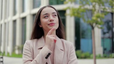 Şık ceketli düşünceli bir kadın dudaklarını büzüp yana bakarken çenesine işaret parmağıyla dokunuyor. Oyuncu tasarımcı modern binaların dış görünüşünü kontrol ediyor ve zihninde yenileme fikirleri yaratıyor..