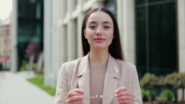 Gözlük takıp kameraya bakarken gülümseyen bej takım elbiseli çekici bir kadının portresi. Profesyonel kadın çalışan, teneffüste dışarıda vakit geçirirken kendinden emin hissediyor..