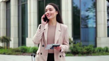 Modern binanın arka planında modern akıllı telefondan konuşurken elinde bilet ve pasaport tutan gülümseyen beyaz kadın. Kadın valizin yanında duruyor ve yurtdışına seyahate hazırlanıyor..