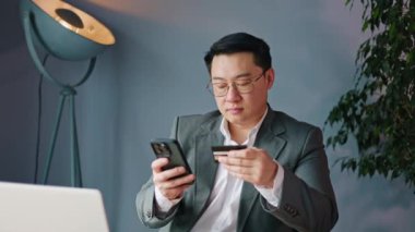 Modern akıllı telefondan alışveriş yaptıktan sonra iş yerinde oturup elinde kredi kartı tutarak hata yapan Japon adam hoşnutsuzdur. Sinirli bir çalışan ödemede sorun yaşıyor..