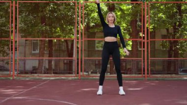 Spor kıyafetli genç bir kadın basketbol sahasında kollarını açıp antrenmanına hazırlanıyor..