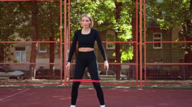 Spor kıyafetleri içinde spor giyinen spor ayakkabılı bir kadın spor sahasındaki bir direniş grubuyla çömelme ve yan bacak kaldırıyor..