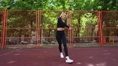 Kısa sarı saçlı genç bir kadın kırmızı spor sahasında siyah spor kıyafetleriyle dans ediyor. Ağaçlar ve turuncu çitlerle çevrili..