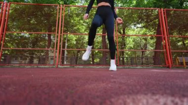 Siyah taytlı ve beyaz ayakkabılı bir kadın bacaklarını ısıtıyor ve sahada ayak hareketlerini çalışıyor..