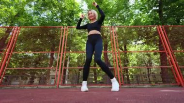 Siyah spor kıyafetleri giyen genç bir kadın güneşli bir günde açık hava spor sahasında dans ediyor ve esniyor..