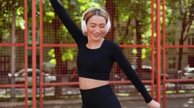 Genç ve mutlu bir bayan dans eder ve kulaklıklarıyla dışarıda siyah bir spor kıyafetiyle müzik dinlemekten hoşlanır.