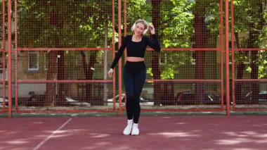 Spor kıyafetleri içinde mutlu bir kadın dışarıda basketbol sahasında kulaklıkla müzik eşliğinde dans eder..