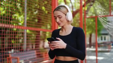 Kulaklık takan genç bir kadın açık hava basketbol sahasında telefonuna bakarken gülümsüyor..