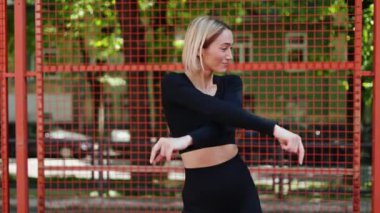 Siyah spor bluzlu ve taytlı mutlu bir genç kadın spor sahasında egzersiz yaparak kaslarını ısıtıyor..
