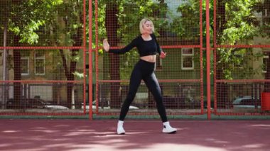 Genç bir kadın, yerel bir basketbol sahasında spor kıyafetleriyle gün ışığında dans ediyor..