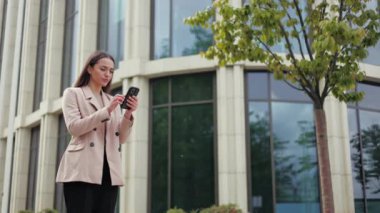 Kafası karışmış beyaz bir kadın, açık havada çağdaş akıllı telefondan sabit internet bağlantısı kurmaya çalışıyor. Modern binanın arka planına karşı memnuniyetsizliğini ifade eden hayal kırıklığına uğramış genç bayan..