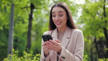 Heyecanlı kadın elinde cep telefonu ekranına bakıyor ve dışarıda dururken zafer jesti yapıyor. Beyaz kadın online bankacılık başvurusundan tebligat alırken seviniyor.