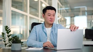 Portatif bilgisayar açıp modern ofiste gülümseyen Çinli girişimci masaüstünde oturuyor. Rahat iş adamı kişisel ekipmanlı kabinede yeni iş gününe hazırlanıyor..