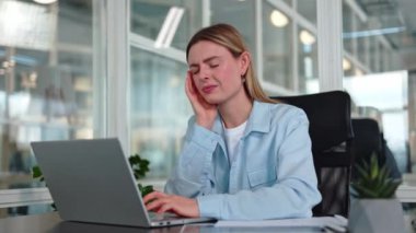 İş yerinde ağza dokunan acı dolu yüz ifadesi olan üzgün, beyaz bir menajer kadın. İş sırasında dizüstü bilgisayarda diş ağrısı çeken, sıradan giyinmiş, hayal kırıklığına uğramış sarışın bir kadın..