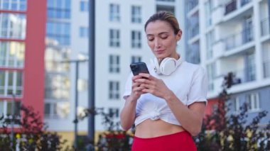 Açık havada egzersiz yaparken kişisel akıllı telefon kullanan gülümseyen genç bir kadın. Neşeli beyaz kadın, modern aletlerin arasında kablosuz kulaklıkla, temiz havada boynuna doğru kayıyor..