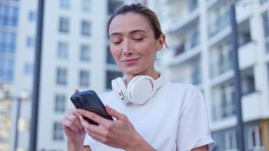 Boynunda kablosuz kulaklıkla dışarıda akıllı telefonlara bakan neşeli beyaz kadın portresi. Beyaz tişört giymiş, gülümseyen genç bir kadın kameraya geniş bir gülümsemeyle bakıyor..
