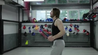 Tarz sahibi, şık saç kesimi olan, kardiyo egzersizleri için fitness ekipmanı kullanan ve nefes almaya devam eden bir erkek. Atletik adam farklı atlama türlerini boş spor salonunda ip atlamakla karıştırıyor..
